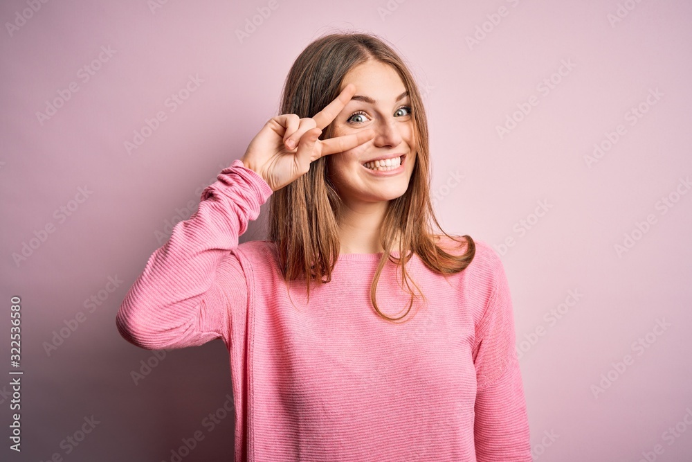 Young beautiful redhead woman wearing casual sweater over isolated pink background Doing peace symbol with fingers over face, smiling cheerful showing victory Young beautiful redhead woman wearing casual sweater over isolated pink background Doing peace symbol with fingers over face, smiling cheerful showing victory