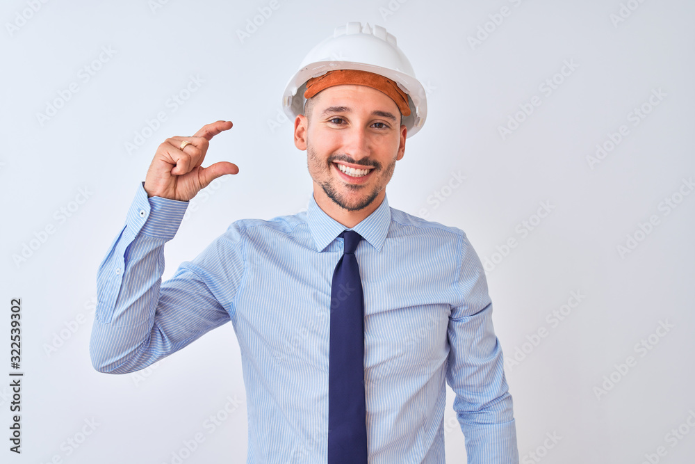 Young business man wearing contractor safety helmet over isolated background smiling and confident gesturing with hand doing small size sign with fingers looking and the camera. Measure concept.