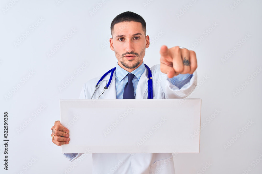 Young doctor man holding blank advertising banner over isolated background pointing with finger to the camera and to you, hand sign, positive and confident gesture from the front Young doctor man holding blank advertising banner over isolated background pointing with finger to the camera and to you, hand sign, positive and confident gesture from the front