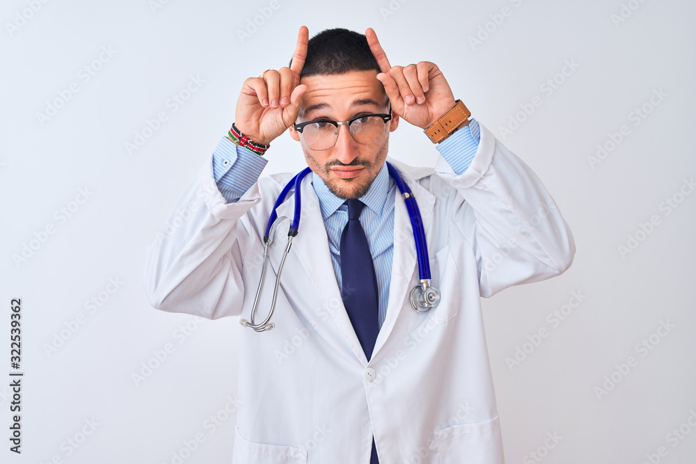 Young doctor man wearing stethoscope over isolated background doing funny gesture with finger over head as bull horns Young doctor man wearing stethoscope over isolated background doing funny gesture with finger over head as bull horns