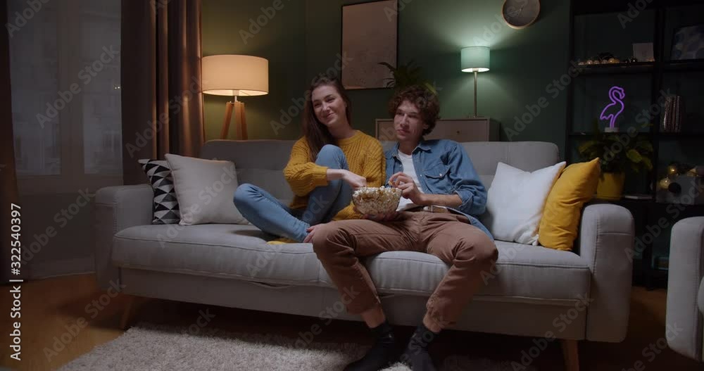 Caucasian happy boyfriend and girlfriend sitting on the sofa at home in the evening with popcorn and watching comedy film on TV. Caucasian happy boyfriend and girlfriend sitting on the sofa at home in the evening with popcorn and watching comedy film on TV.