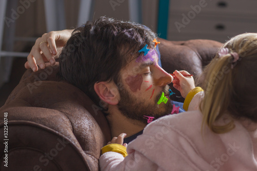 Картината върху платно Young father sleeping on the couch while his little daughter paints his face wit