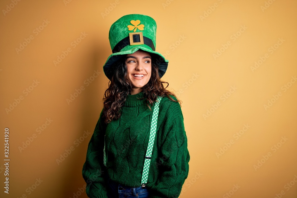 Beautiful curly hair woman wearing green hat with clover celebrating saint patricks day looking away to side with smile on face, natural expression. Laughing confident.