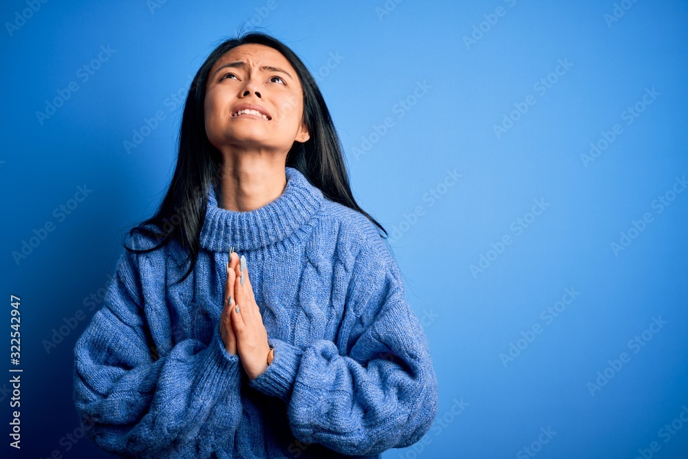 Young beautiful chinese woman wearing casual sweater over isolated blue background begging and praying with hands together with hope expression on face very emotional and worried. Begging. Young beautiful chinese woman wearing casual sweater over isolated blue background begging and praying with hands together with hope expression on face very emotional and worried. Begging.
