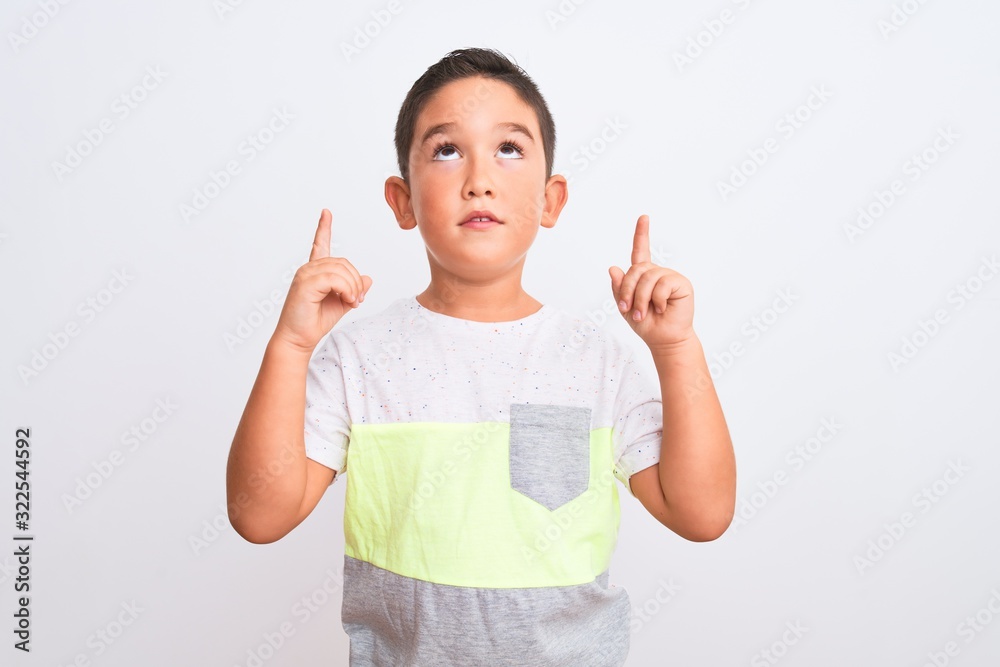 Beautiful kid boy wearing casual t-shirt standing over isolated white background amazed and surprised looking up and pointing with fingers and raised arms. Beautiful kid boy wearing casual t-shirt standing over isolated white background amazed and surprised looking up and pointing with fingers and raised arms.