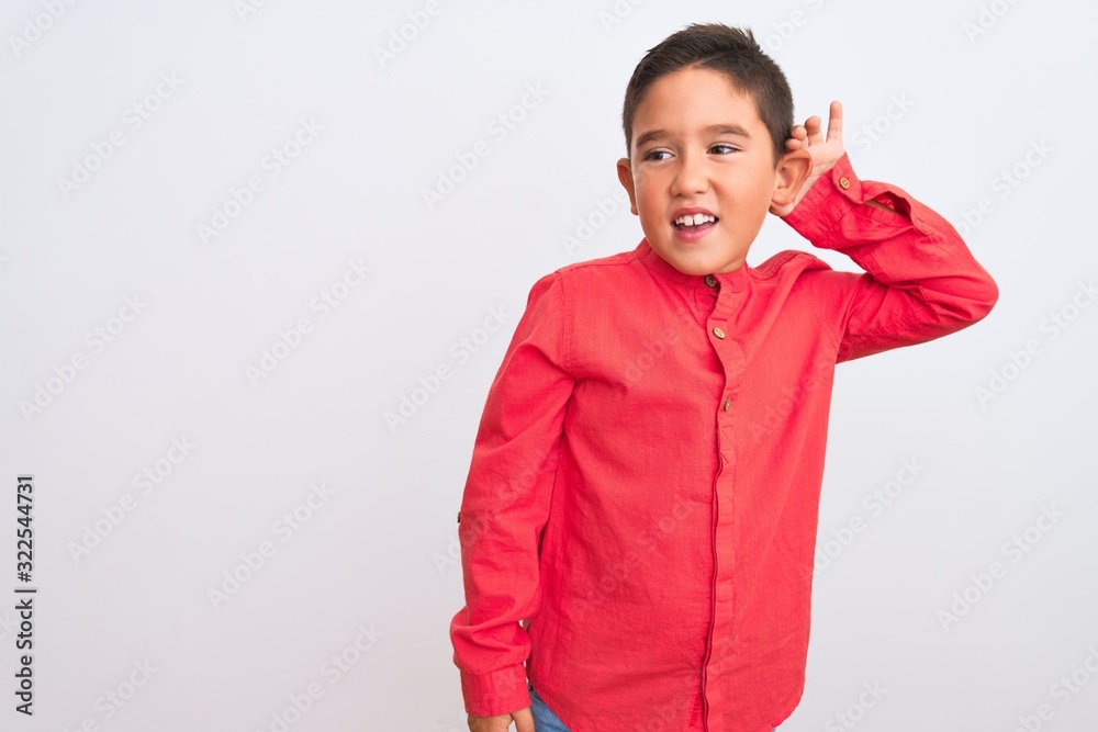 Beautiful kid boy wearing elegant red shirt standing over isolated white background smiling with hand over ear listening an hearing to rumor or gossip. Deafness concept. Beautiful kid boy wearing elegant red shirt standing over isolated white background smiling with hand over ear listening an hearing to rumor or gossip. Deafness concept.