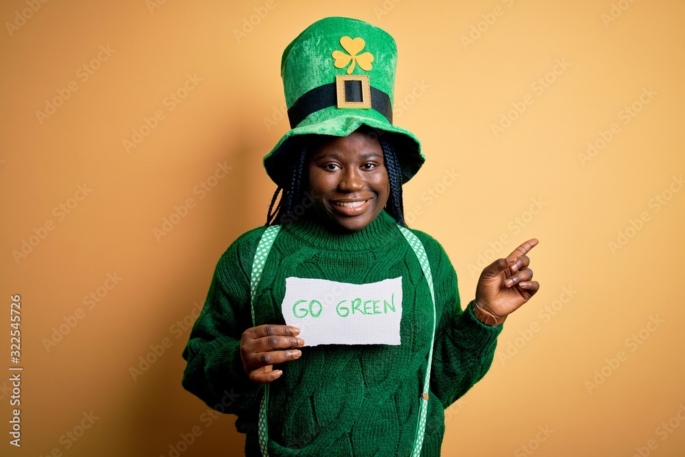 Plus size african american woman wearing green hat holding paper on saint patricks day very happy pointing with hand and finger to the side