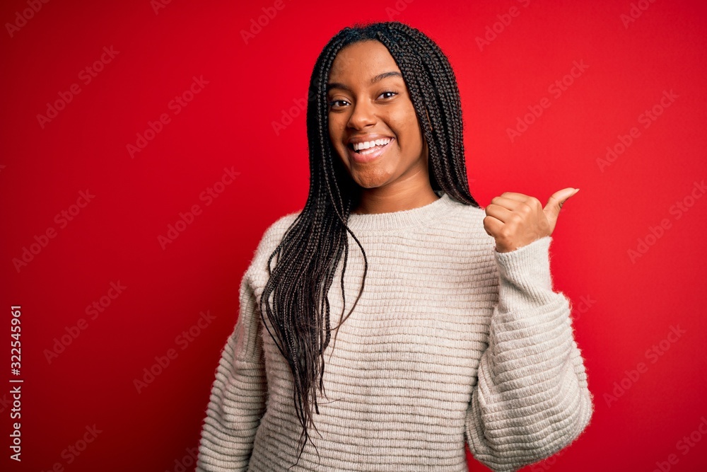 Young african american woman wearing casual winter sweater over red isolated background smiling with happy face looking and pointing to the side with thumb up. Young african american woman wearing casual winter sweater over red isolated background smiling with happy face looking and pointing to the side with thumb up.