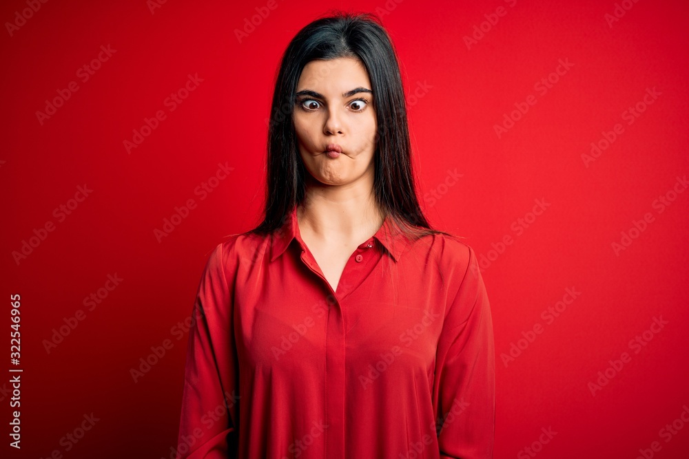 Young beautiful brunette woman wearing casual shirt standing over red background making fish face with lips, crazy and comical gesture. Funny expression. Young beautiful brunette woman wearing casual shirt standing over red background making fish face with lips, crazy and comical gesture. Funny expression.