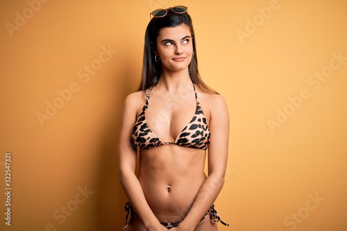 Young beautiful brunette woman on vacation wearing swimwear bikini over yellow background smiling looking to the side and staring away thinking.