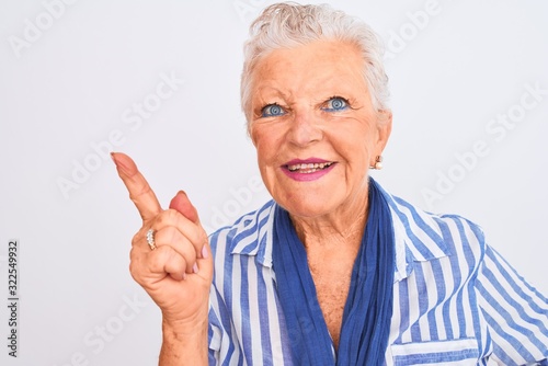 Senior grey-haired woman wearing blue striped shirt standing over isolated white background very happy pointing with hand and finger to the side