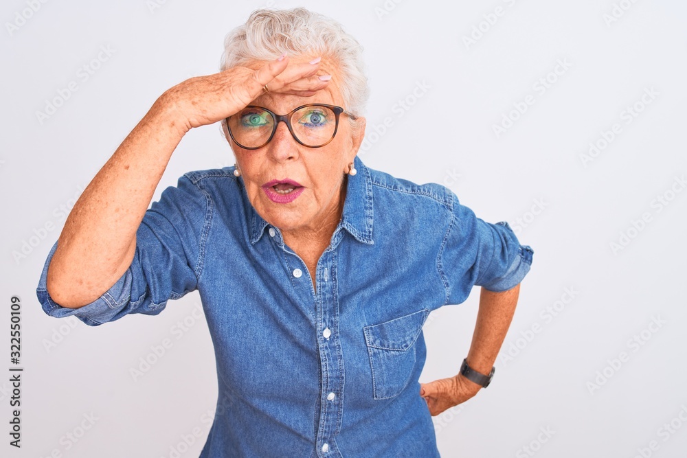 Senior grey-haired woman wearing denim shirt and glasses over isolated white background very happy and smiling looking far away with hand over head. Searching concept.