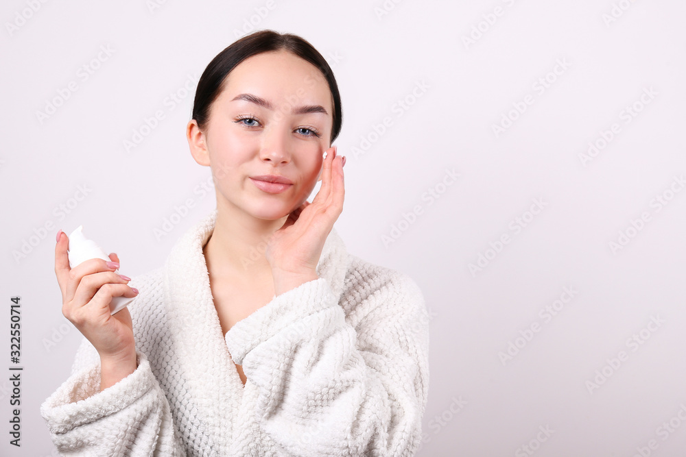 Portrait of young beautiful woman with perfectly clean face skin. Female with long black hair tied in ponytail wearing bathrobe, applying moisturizing cream. Close up, copy space background. Portrait of young beautiful woman with perfectly clean face skin. Female with long black hair tied in ponytail wearing bathrobe, applying moisturizing cream. Close up, copy space background.