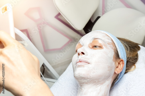 Young adult woman lying on bed at beauty spa salon with applied facial cosmetic mask making phone selfie shot for social networks. Influencer making online vlog stream with skin care product review