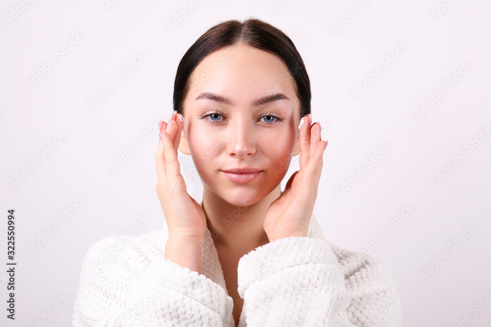 Portrait of young beautiful woman with perfectly clean face skin. Female with long black hair tied in ponytail wearing bathrobe, applying moisturizing cream. Close up, copy space background. Portrait of young beautiful woman with perfectly clean face skin. Female with long black hair tied in ponytail wearing bathrobe, applying moisturizing cream. Close up, copy space background.