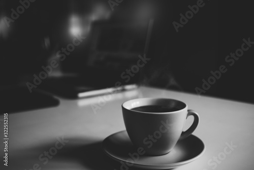 The monochrome tone of aroma black coffee on cup with a smoke on the table, laptop office at home