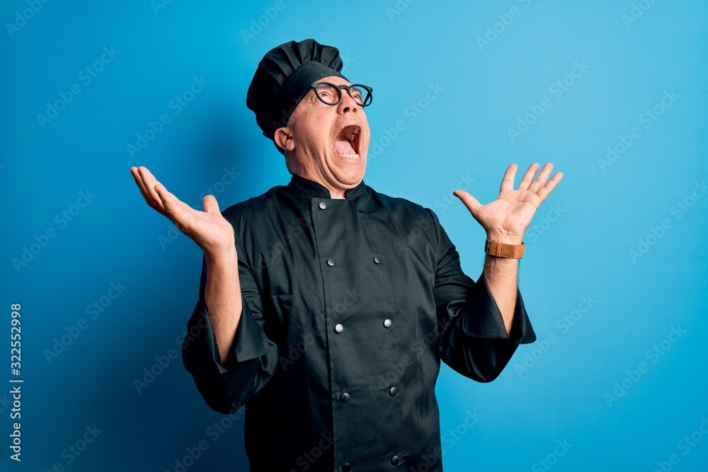 Middle age handsome grey-haired chef man wearing cooker uniform and hat crazy and mad shouting and yelling with aggressive expression and arms raised. Frustration concept. Middle age handsome grey-haired chef man wearing cooker uniform and hat crazy and mad shouting and yelling with aggressive expression and arms raised. Frustration concept.