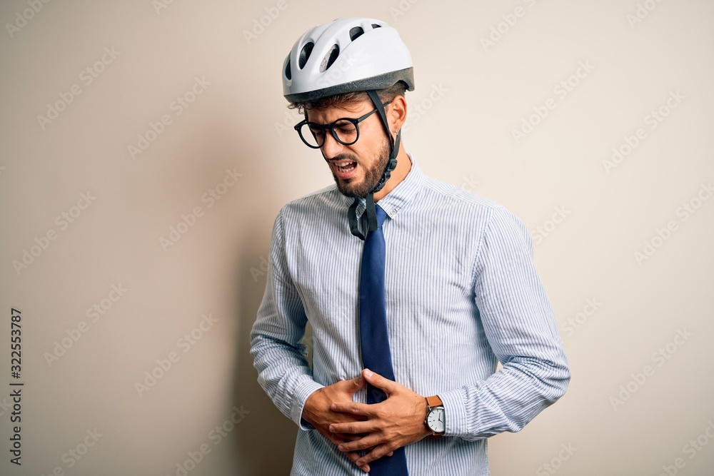 Young businessman wearing glasses and bike helmet standing over isolated white bakground with hand on stomach because nausea, painful disease feeling unwell. Ache concept. Young businessman wearing glasses and bike helmet standing over isolated white bakground with hand on stomach because nausea, painful disease feeling unwell. Ache concept.