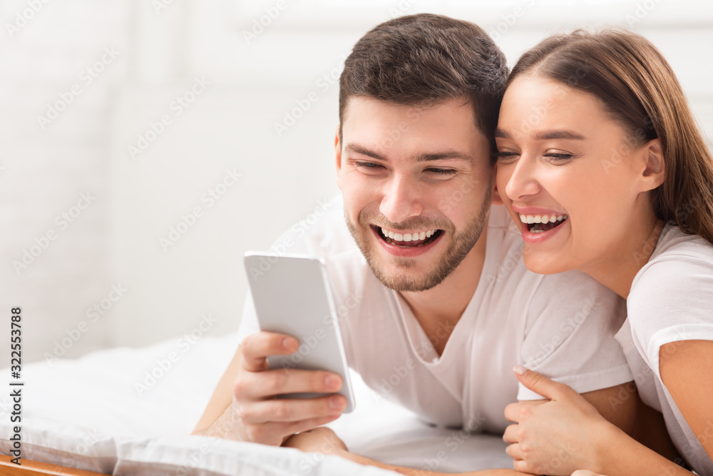 Happy Couple Using Smartphone Texting Lying In Bed In Bedroom