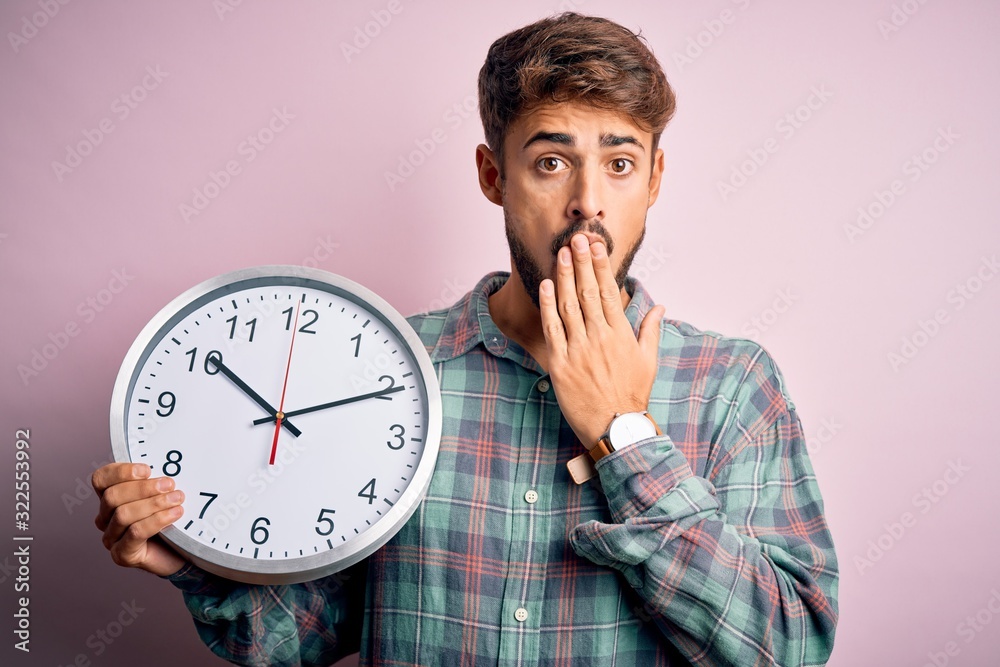 Young man with beard doing countdown using big clock over isolated pink background cover mouth with hand shocked with shame for mistake, expression of fear, scared in silence, secret concept Young man with beard doing countdown using big clock over isolated pink background cover mouth with hand shocked with shame for mistake, expression of fear, scared in silence, secret concept