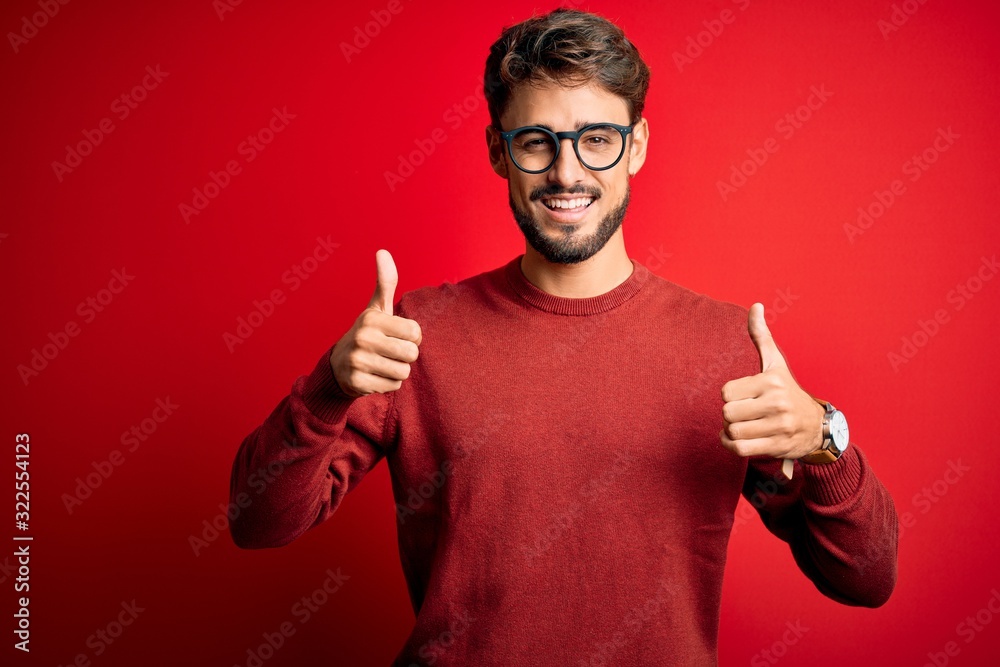 Young handsome man with beard wearing glasses and sweater standing over red background success sign doing positive gesture with hand, thumbs up smiling and happy. Cheerful expression Young handsome man with beard wearing glasses and sweater standing over red background success sign doing positive gesture with hand, thumbs up smiling and happy. Cheerful expression