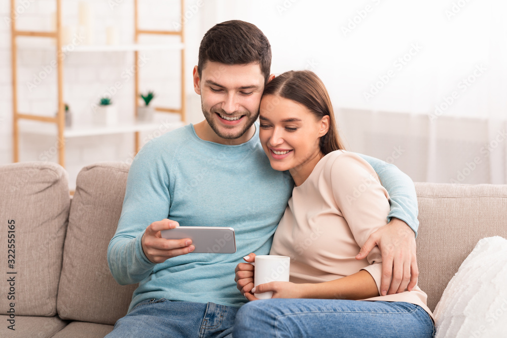 Happy couple using smartphone networking sitting on sofa indoor Happy couple using smartphone networking sitting on sofa indoor