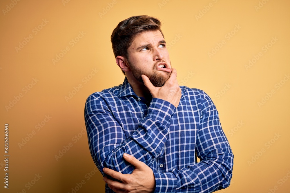 Young blond businessman with beard and blue eyes wearing shirt over yellow background Thinking worried about a question, concerned and nervous with hand on chin Young blond businessman with beard and blue eyes wearing shirt over yellow background Thinking worried about a question, concerned and nervous with hand on chin