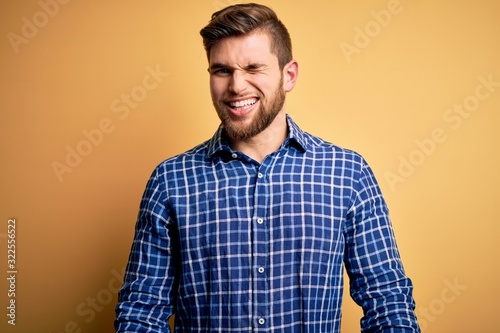 Young blond businessman with beard and blue eyes wearing shirt over yellow background winking looking at the camera with sexy expression, cheerful and happy face.