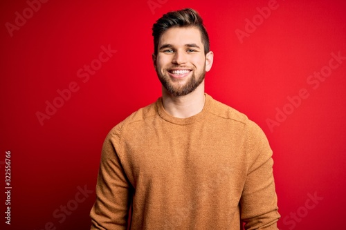 Young blond man with beard and blue eyes wearing casual shirt over red background with a happy and cool smile on face. Lucky person.