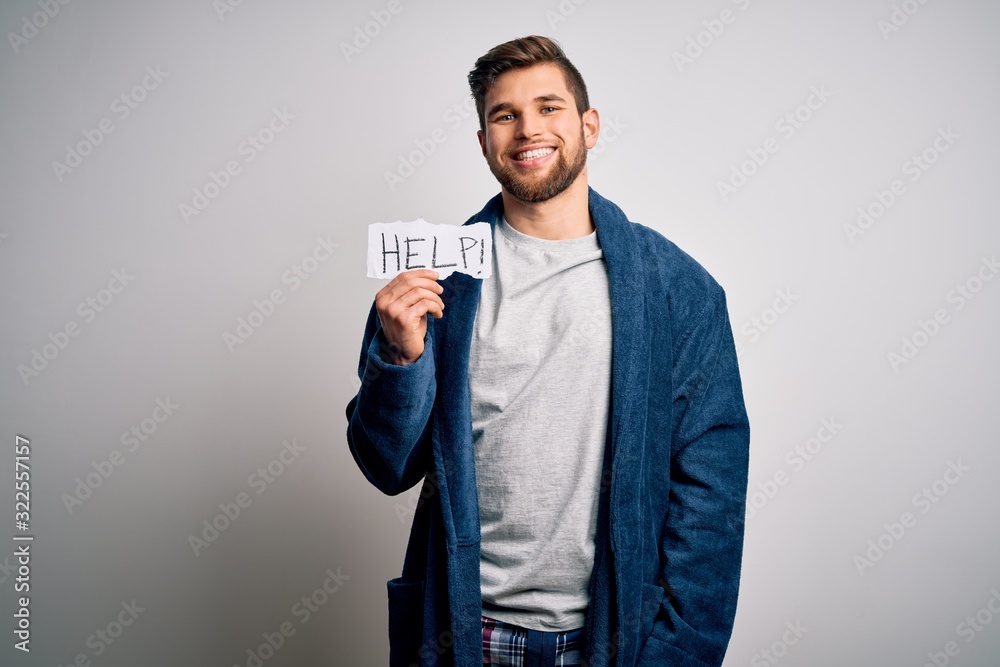 Young blond man with beard and blue eyes wearing pajama holding paper with help message with a happy face standing and smiling with a confident smile showing teeth