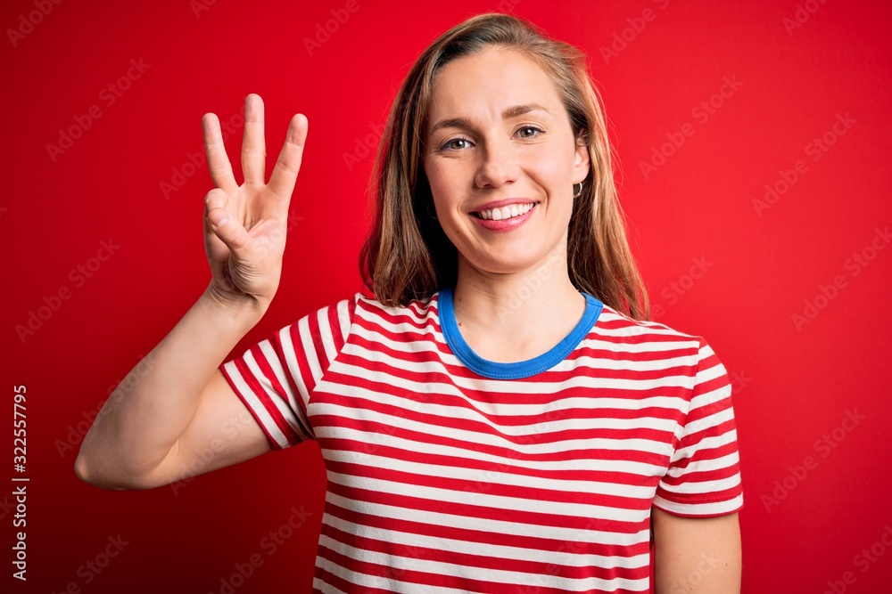 Young beautiful blonde woman wearing casual striped t-shirt over isolated red background showing and pointing up with fingers number three while smiling confident and happy. Young beautiful blonde woman wearing casual striped t-shirt over isolated red background showing and pointing up with fingers number three while smiling confident and happy.
