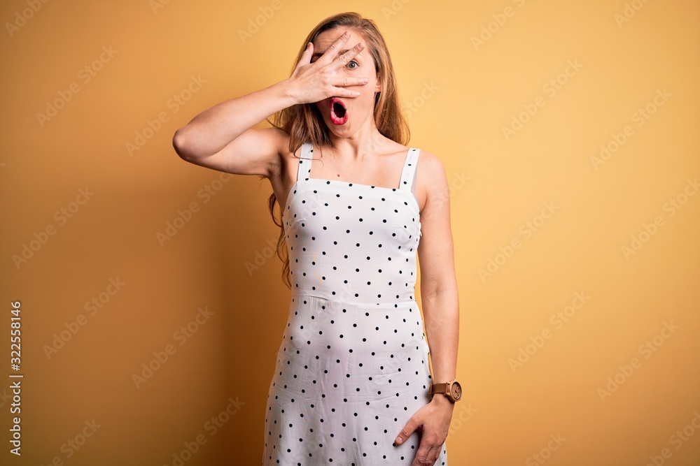 Young beautiful blonde woman on vacation wearing summer dress over yellow background peeking in shock covering face and eyes with hand, looking through fingers with embarrassed expression. Young beautiful blonde woman on vacation wearing summer dress over yellow background peeking in shock covering face and eyes with hand, looking through fingers with embarrassed expression.