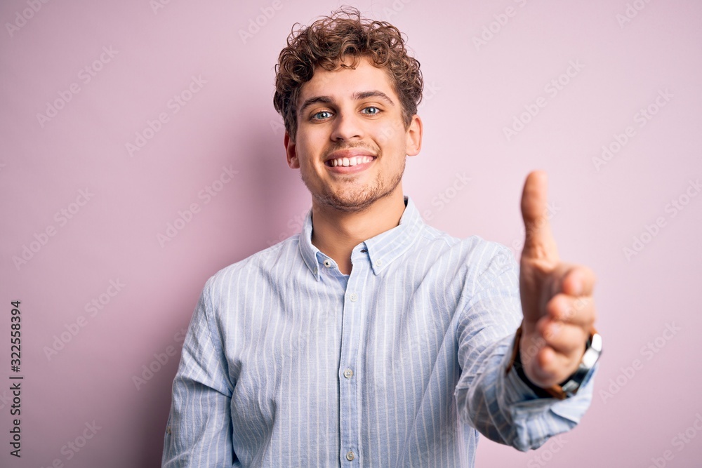 Young blond handsome man with curly hair wearing striped shirt over white background smiling friendly offering handshake as greeting and welcoming. Successful business. Young blond handsome man with curly hair wearing striped shirt over white background smiling friendly offering handshake as greeting and welcoming. Successful business.