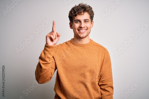 Young blond handsome man with curly hair wearing casual sweater over white background showing and pointing up with finger number one while smiling confident and happy.