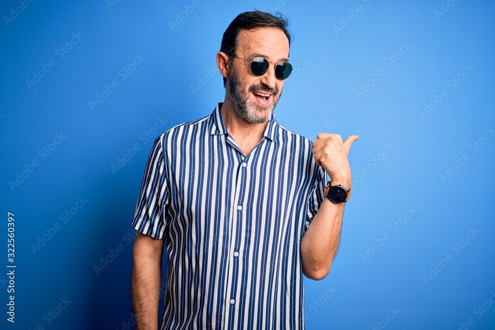 Naklejka premium Middle age hoary man wearing striped shirt and sunglasses over isolated blue background smiling with happy face looking and pointing to the side with thumb up.