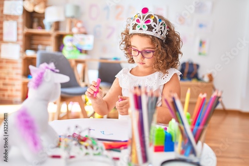 Beautiful toddler wearing glasses and princess crown sitting drawing using paper and marker pen at kindergarten