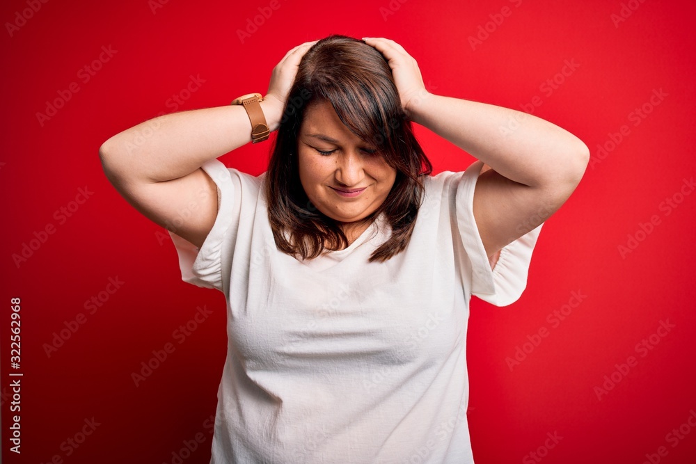 Beautiful brunette plus size woman wearing casual t-shirt over isolated red background suffering from headache desperate and stressed because pain and migraine. Hands on head. Beautiful brunette plus size woman wearing casual t-shirt over isolated red background suffering from headache desperate and stressed because pain and migraine. Hands on head.