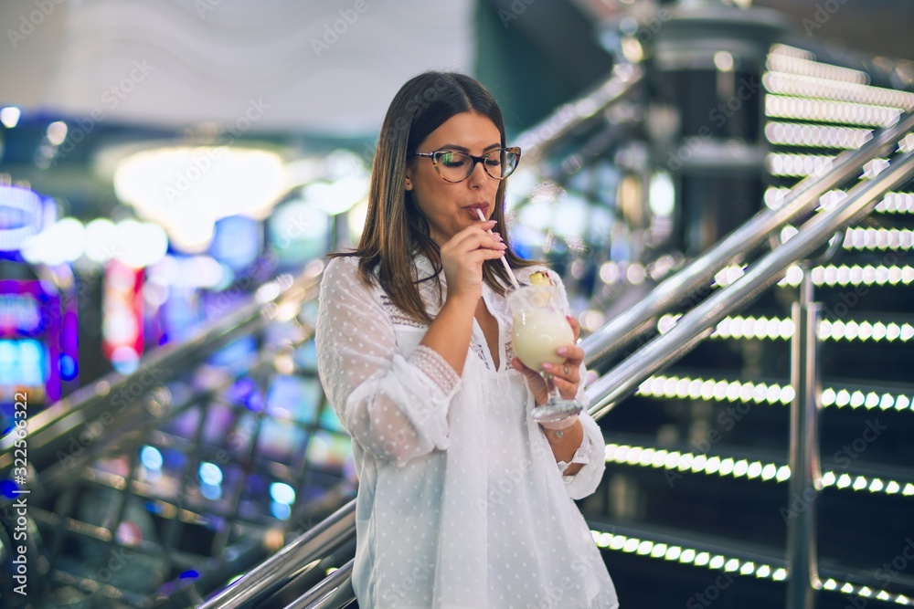 Young beautiful woman on vacation smiling happy and confident. Standing with smile on face drinking cocktail around lots of slot machine at casino