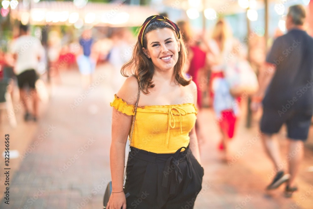Fototapeta premium Young beautiful girl smiling happy and confident walking at the town street, standing with a smile on face around night lights bokeh
