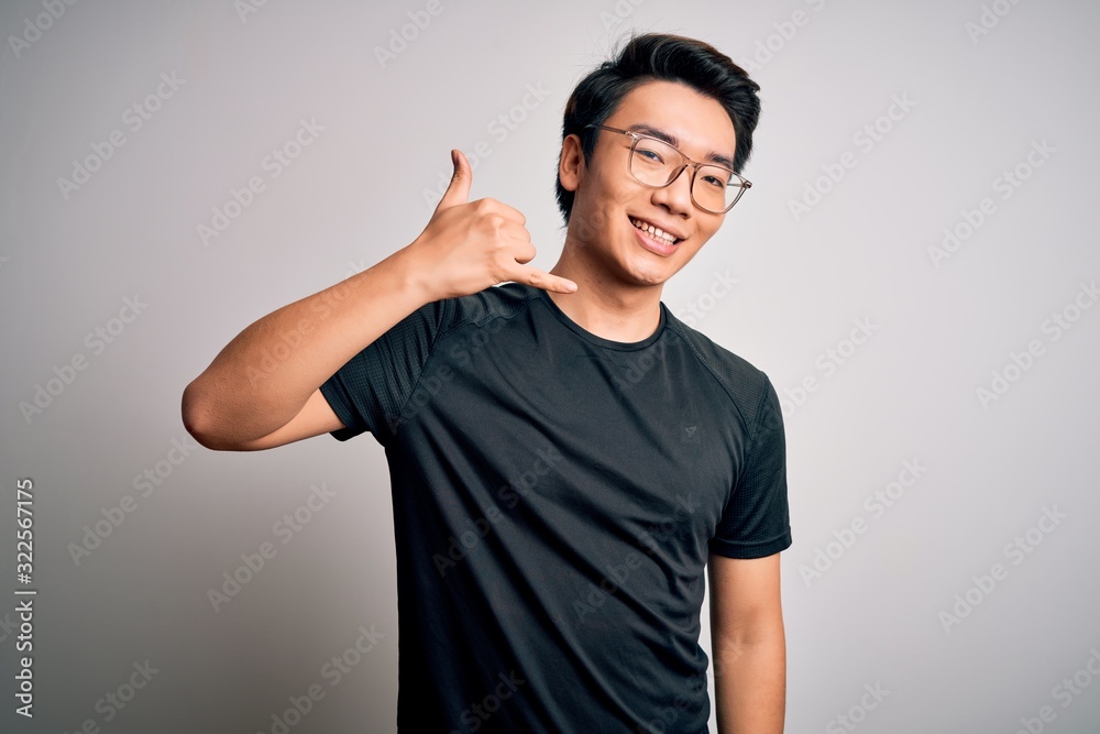 Young handsome chinese man wearing black t-shirt and glasses over white background smiling doing phone gesture with hand and fingers like talking on the telephone. Communicating concepts. Young handsome chinese man wearing black t-shirt and glasses over white background smiling doing phone gesture with hand and fingers like talking on the telephone. Communicating concepts.