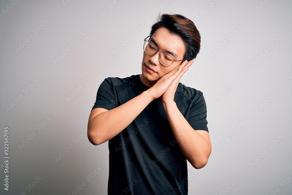 Young handsome chinese man wearing black t-shirt and glasses over white background sleeping tired dreaming and posing with hands together while smiling with closed eyes. Young handsome chinese man wearing black t-shirt and glasses over white background sleeping tired dreaming and posing with hands together while smiling with closed eyes.