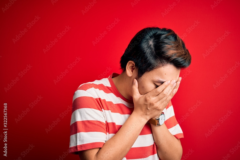 Young handsome chinese man wearing casual striped t-shirt standing over red background with sad expression covering face with hands while crying. Depression concept. Young handsome chinese man wearing casual striped t-shirt standing over red background with sad expression covering face with hands while crying. Depression concept.
