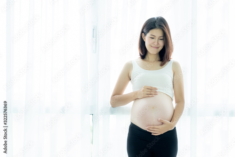 A beautiful Asian woman standing in front of the window and touching her belly. Pregnancy and expectation concept A beautiful Asian woman standing in front of the window and touching her belly. Pregnancy and expectation concept