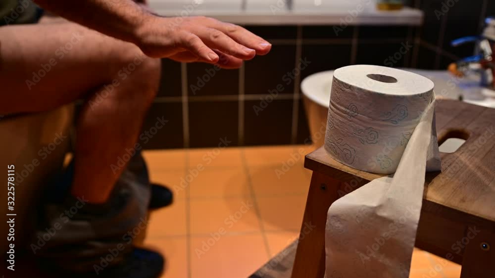 Close-up image of the toilet paper.In the blurred background you can recognize the bare legs of a Caucasian man as he defecates.The right hand tries to grab the toilet paper