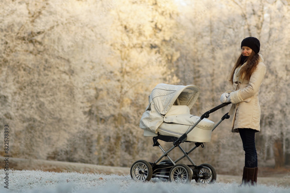 Young woman in white coat standing with white baby stroller in sunny winter park. Childcare and love Young woman in white coat standing with white baby stroller in sunny winter park. Childcare and love
