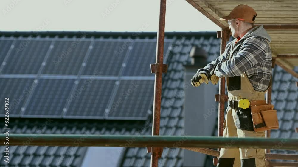 Construction Worker on a Scaffolding Looking Around. House Roof with Solar Panels in a Background. Construction Worker on a Scaffolding Looking Around. House Roof with Solar Panels in a Background.