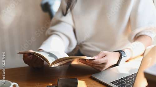 Cropped image of young beautiful woman holding/reading a books in her hands in front a computer laptop, coffee cup and smartphone at the modern wooden table with comfortable living room as background. photo