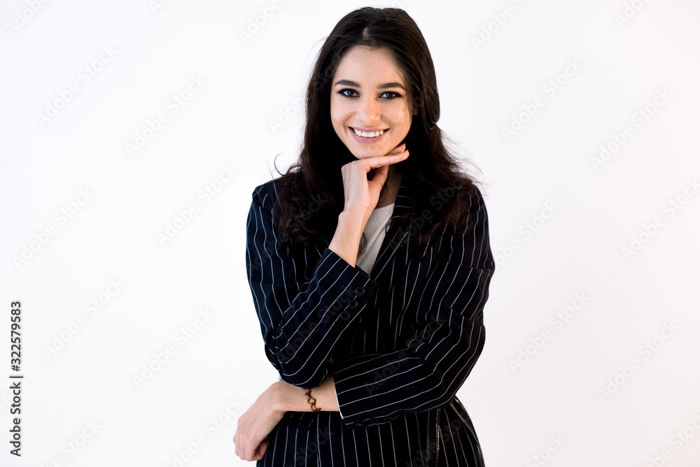 Smiling pretty Caucasian business woman in black jacket with one hand touching chin, standing against white background. Toothy smile, horizontal shot. Smiling pretty Caucasian business woman in black jacket with one hand touching chin, standing against white background. Toothy smile, horizontal shot.