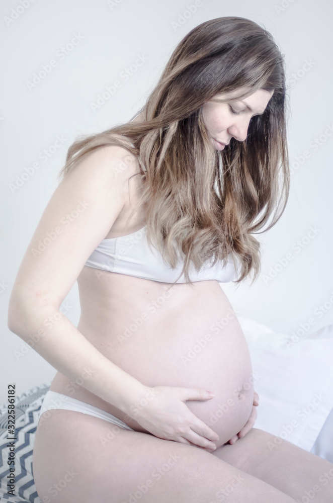 Mujer embarazada de perfil derecho en ropa interior en la cama con fondo blanco Mujer embarazada de perfil derecho en ropa interior en la cama con fondo blanco