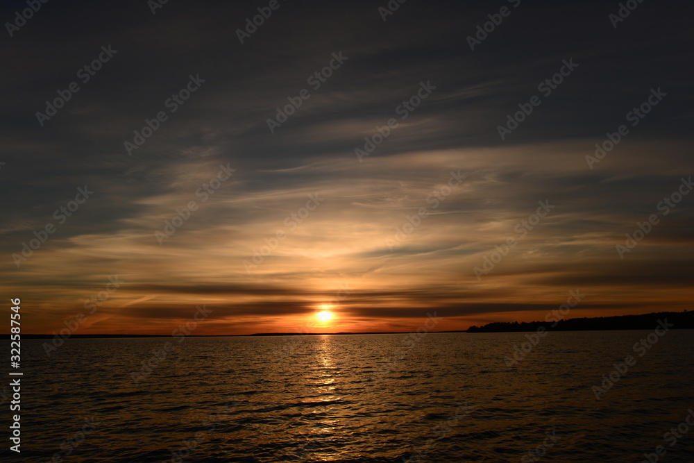 Spring bright sun glowing at sunset in the clouds of blue sky on the horizon above the lake water